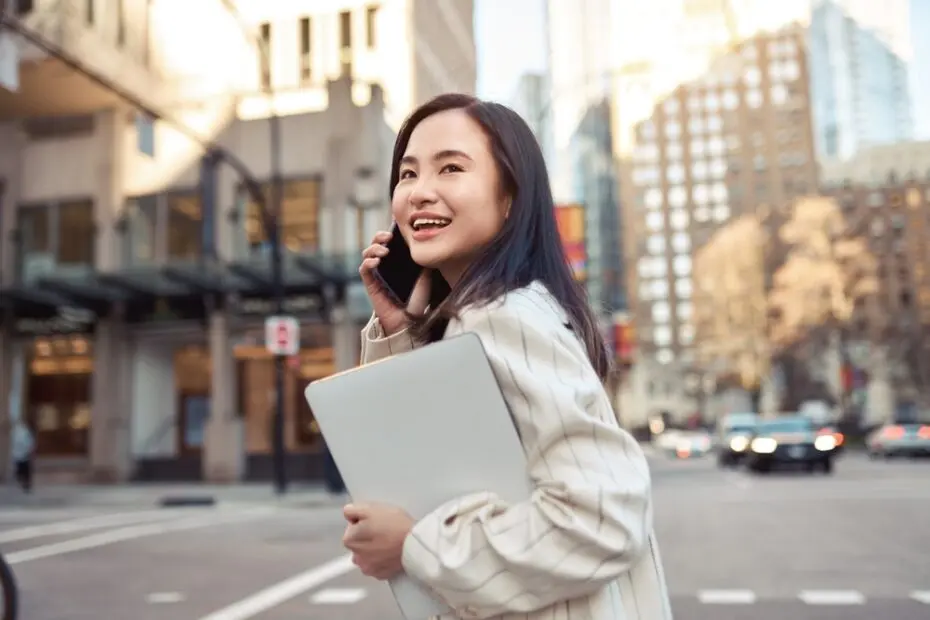 Young busy happy Asian business woman office professional holding cellphone in hands walking on big city urban street making corporate business call, talking on the cellular phone.